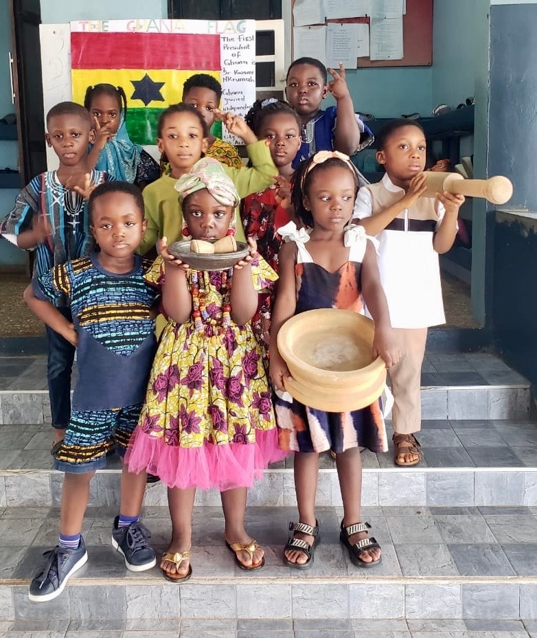 Students in traditional dress holding a mortar and pestle in front of the Ghana flag